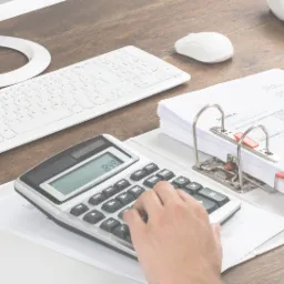 Osoba w białej koszuli, siedząca przy biurku z drewnianym blatem, wpisuje dane na kalkulatorze obok segregatora z dokumentami, z widocznymi elementami biurowymi jak monitor, klawiatura, myszka...