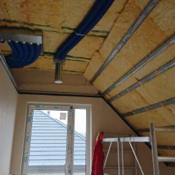 Izolacja poddasza wełną mineralną z widocznymi niebieskimi rurami wentylacyjnymi i metalową konstrukcją sufitu podwieszanego, widok z dołu na okno i rusztowanie.