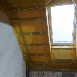 Poddasze w trakcie izolacji, widoczne ocieplenie wełną mineralną pod folią paroizolacyjną, drewniany stelaż i okno dachowe.