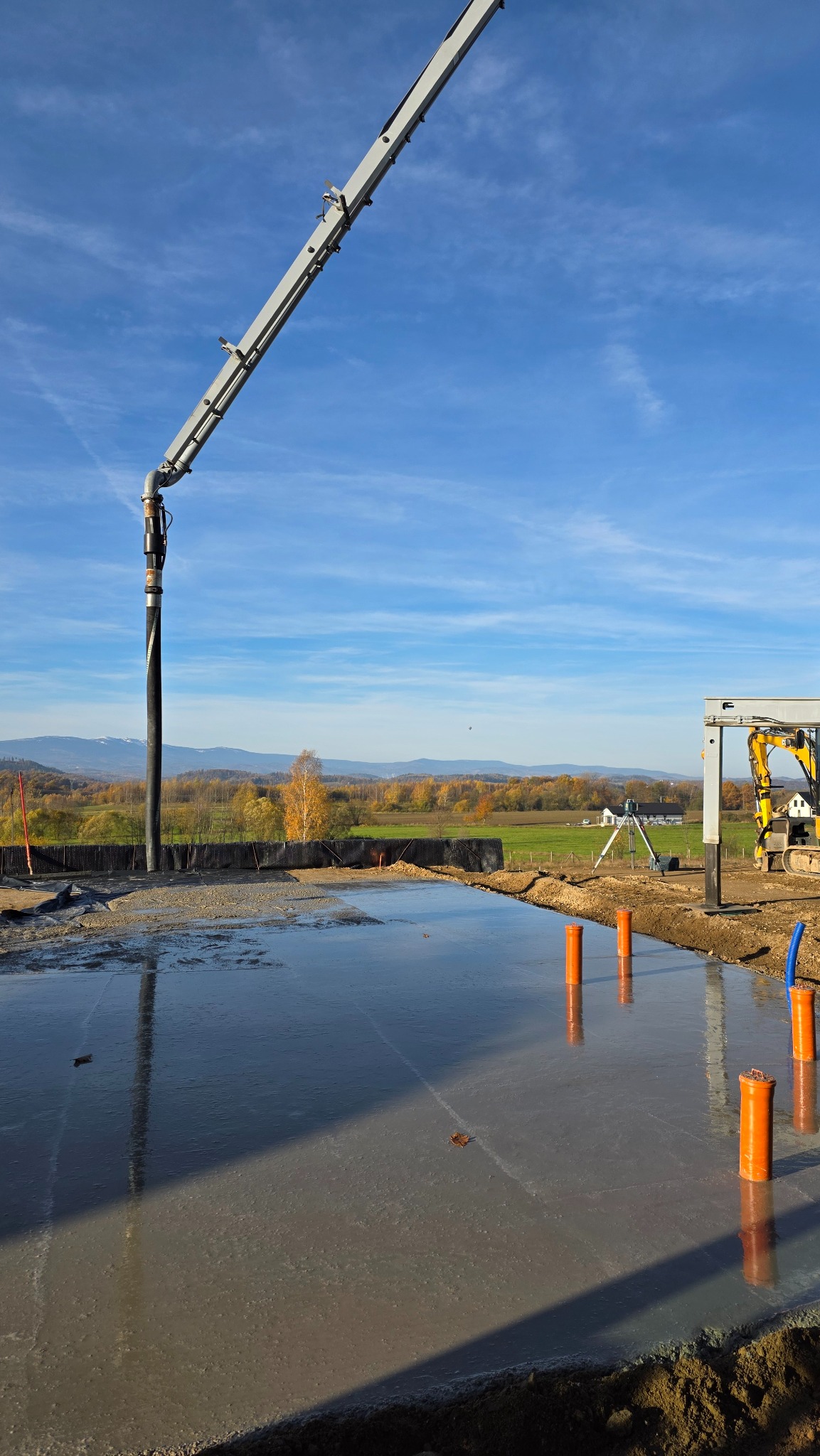 Wylewka betonowa na budowie domu jednorodzinnego z widoczną pompą do betonu, pomarańczowymi rurami i żółtą koparką w tle, przy słonecznej pogodzie.