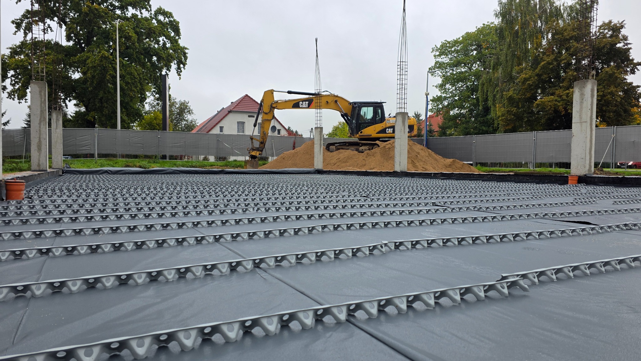Fundament domu z szalunkiem traconym i wykopem ziemi, na którym stoi koparka CAT. Widoczne zbrojenia i ogrodzenie terenu budowy. Pochmurny dzień.