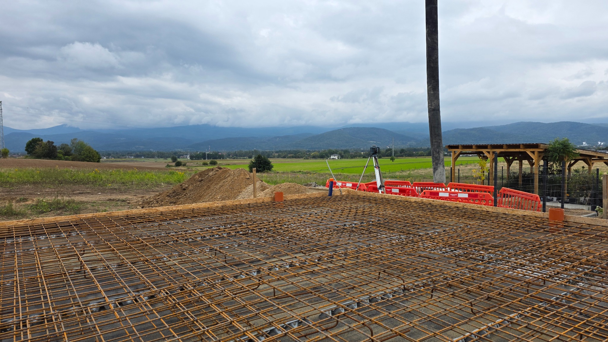 Zbrojenie fundamentów domu jednorodzinnego na tle krajobrazu z górami i pochmurnym niebem. Widoczne pomarańczowe barierki i stelaż pod wylewkę.