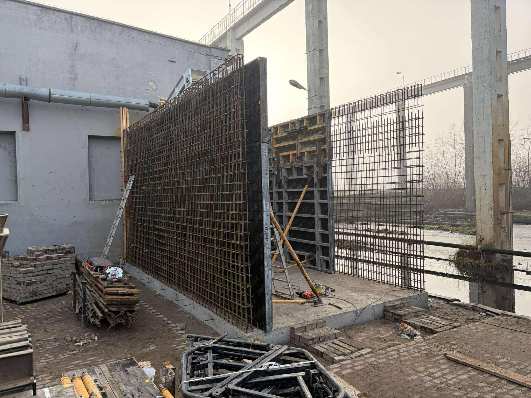 Konstrukcja zbrojeniowa z siatki stalowej i szalunków na budowie, widok z boku, z narzędziami i materiałami budowlanymi w tle, pochmurny dzień.