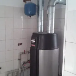 Zamontowany piec grzewczy z podłączonymi rurami, zbiornikiem wyrównawczym i instalacją hydrauliczną w pomieszczeniu wykończonym białymi płytkami.