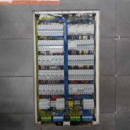 Rozdzielnica elektryczna z licznymi zabezpieczeniami i okablowaniem, zamontowana w ścianie wyłożonej szarą płytką ceramiczną, widoczne fugi i pomarańczowe ślady po taśmie malarskiej.