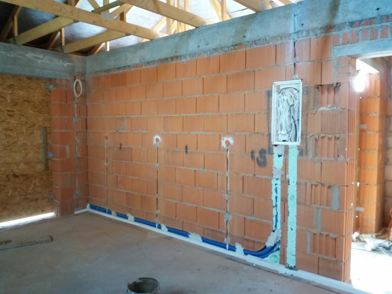 Wnętrze w trakcie budowy z widoczną instalacją elektryczną: kable w peszlach prowadzone w bruzdach w ścianie z cegły, puszki pod gniazdka i otwarta skrzynka rozdzielcza.