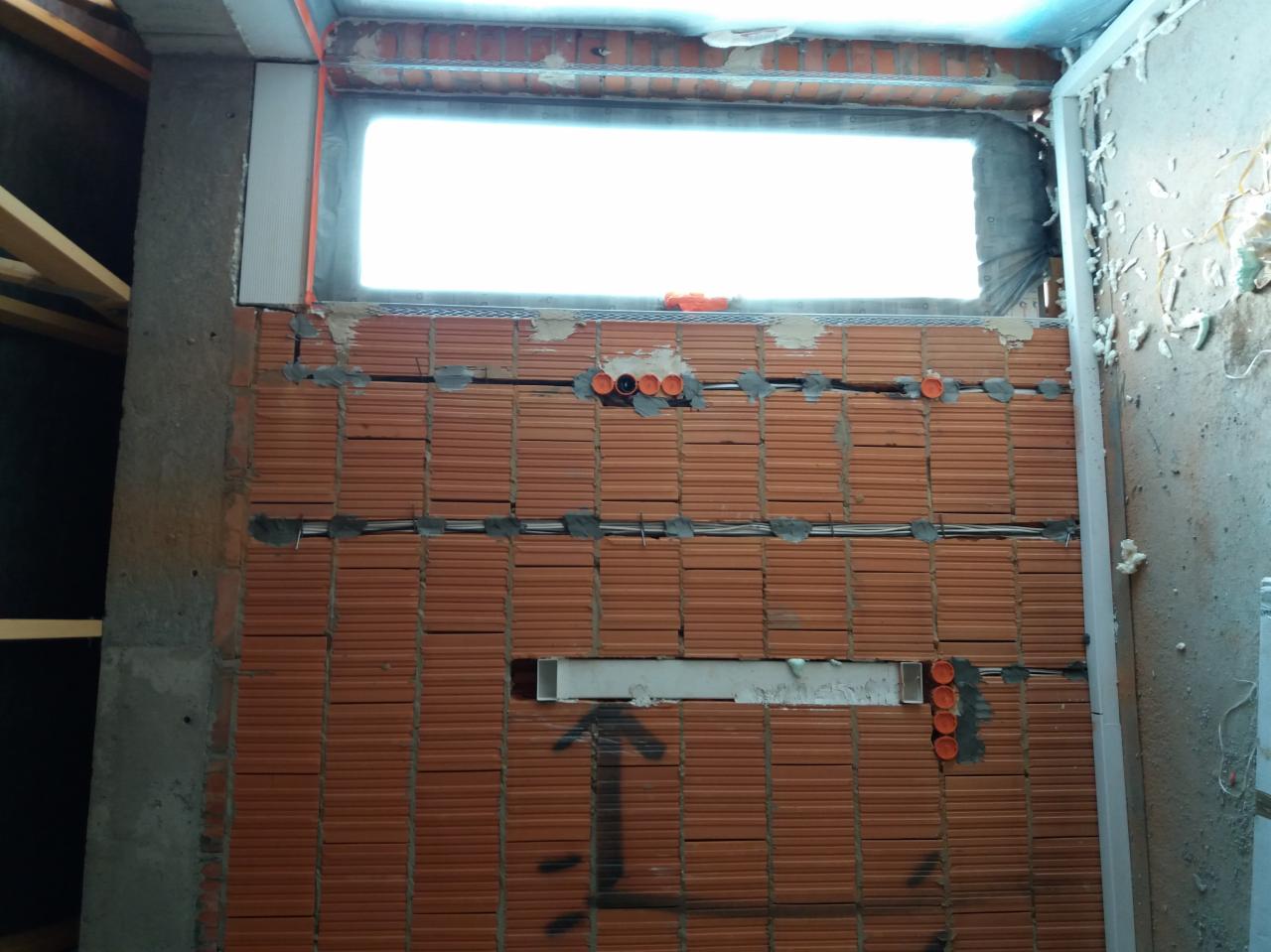Ściana z cegły z widocznymi przewodami elektrycznymi w trakcie montażu, pomarańczowe puszki instalacyjne osadzone w murze, betonowy filar z lewej strony.