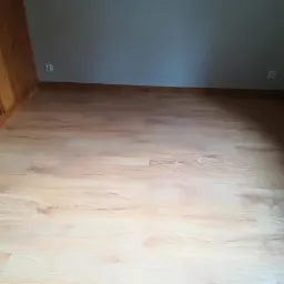 Jasne panele podłogowe z widoczną strukturą drewna, ułożone w pokoju z szarą ścianą i drewnianą boazerią, widoczne gniazdka elektryczne.