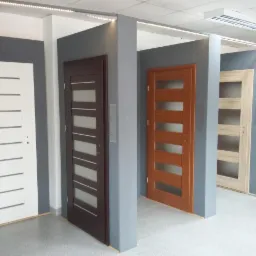 Ekspozycja czterech nowoczesnych drzwi wewnętrznych z przeszkleniami, w kolorach białym, brązowym, miodowym i jasnego drewna, umieszczonych w szarych wnękach w salonie sprzedaży.