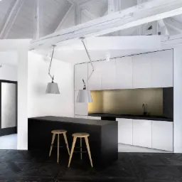 Wnętrze domu z aneksem kuchennym w minimalistycznym stylu. Czarny parkiet w jodełkę, białe ściany i sufit z drewnianymi belkami. Na pierwszym planie czarny barek z dwoma drewnianymi stołkami. W tle...