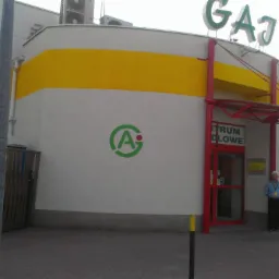 Fasada budynku handlowego z żółtym pasem i zielonym logo, wejście z czerwonymi ramami i widoczna osoba w niebieskiej koszuli.