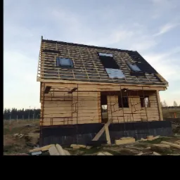 Surowy dom z bali w trakcie budowy, widoczne rusztowania, brak okien i drzwi, częściowo pokryty dach z oknami dachowymi, teren budowy.