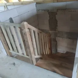 Surowe drewniane schody z balustradą w trakcie montażu w surowym wnętrzu z białych bloczków, widoczne niedoskonałości i brak wykończenia.