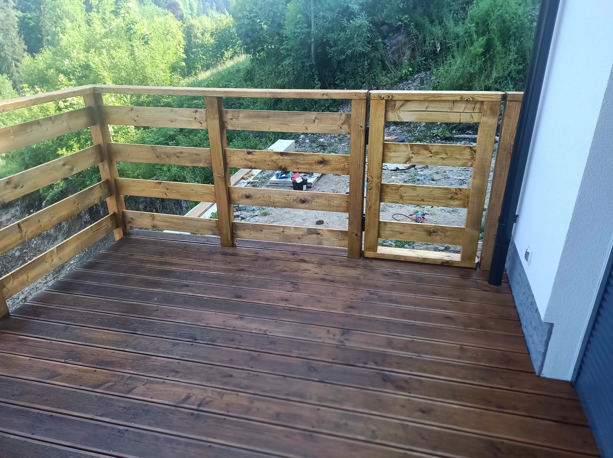 Drewniany taras z balustradą i furtką, widok z góry. Deski tarasowe w odcieniach brązu, drewniana balustrada w naturalnym kolorze drewna. W tle zieleń drzew.