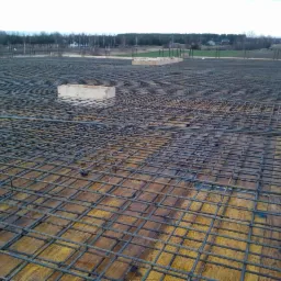 Zbrojenie płyty fundamentowej z widoczną drewnianą skrzynką szalunkową, przygotowane do wylania betonu, na tle krajobrazu z drzewami i polami.