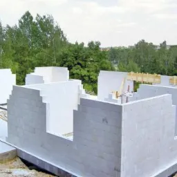 Wznoszenie ścian parterowego budynku z białych bloczków, widoczne drewniane nadproża okienne, w tle drzewa.