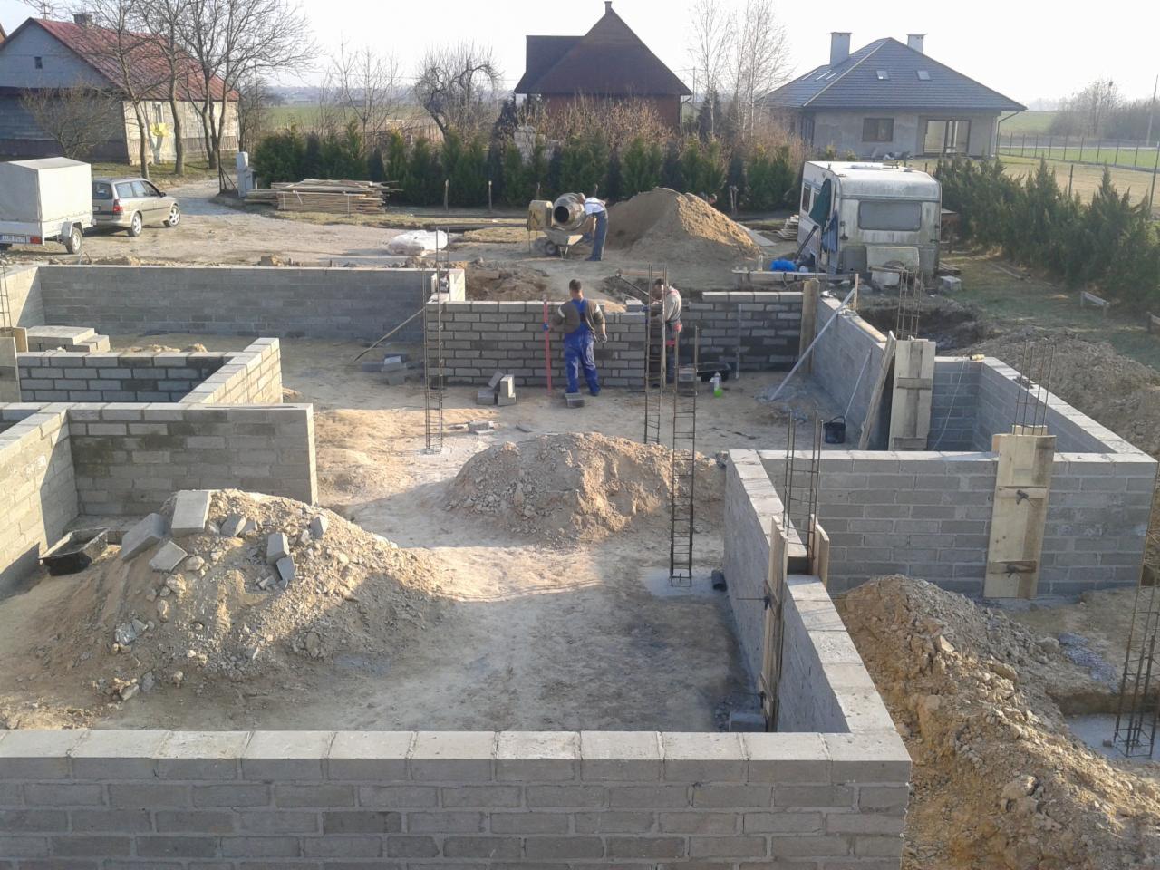 Wznoszenie fundamentów z bloczków betonowych na placu budowy, widoczne zbrojenia, piasek, betoniarka i pracownicy w tle.