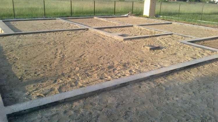 Przygotowany fundament pod budynek z widocznymi zbrojeniami i betonowymi obrzeżami, wypełniony piaskiem.