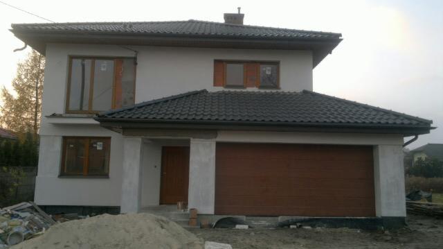 Dwukondygnacyjny dom murowany w trakcie budowy, z szarą dachówką, częściowo otynkowany, z brązową bramą garażową i oknami, widoczny stos piasku i gruzu.