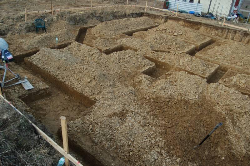 Wykopane rowy pod fundamenty domu z widocznymi deskami szalunkowymi, stertami ziemi i zielonym krzesłem w tle.