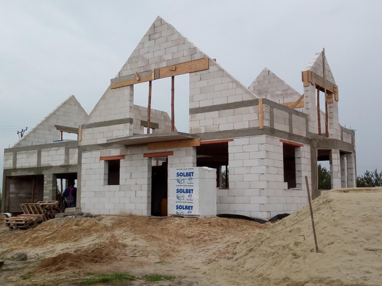 Surowy stan domu z białych bloczków, widoczne nadproża okienne z cegły, palety z pustakami Solbet stojące przed budynkiem, teren budowy z piaskiem.