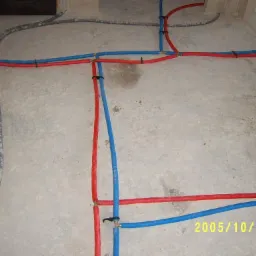 Instalacja wodna i grzewcza w trakcie montażu, rury w kolorach czerwonym i niebieskim ułożone na surowym betonie, widoczne złącza i mocowania.