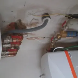 Instalacja grzewcza z widocznym kotłem, rurami miedzianymi z czerwonymi zaworami oraz filtrem siatkowym na tle surowego sufitu z widocznymi przewodami.