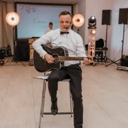 Mężczyzna w muszce grający na czarnej gitarze akustycznej na scenie z nagłośnieniem i oświetleniem podczas wesela.