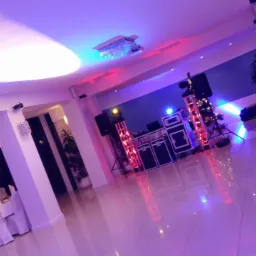 Sala bankietowa przygotowana na uroczystość, widoczne stoły z białymi obrusami, dekoracje świetlne i sprzęt nagłośnieniowy.