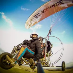 Dwóch mężczyzn w kaskach i kombinezonach leci motoparalotnią z napisem EC EXTREME na tle błękitnego nieba z chmurami i zielonych pól.