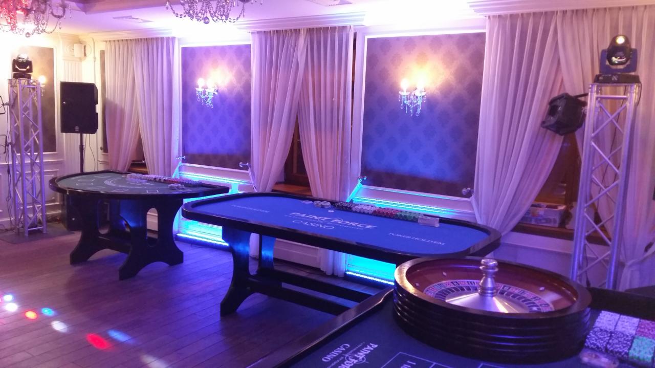 Sala z oświetleniem w kolorze fioletowym, przygotowana do gry w kasyno z stołami do pokera i ruletką, oświetlenie sceniczne na statywach, białe zasłony i dekoracyjne kinkiety na ścianach.