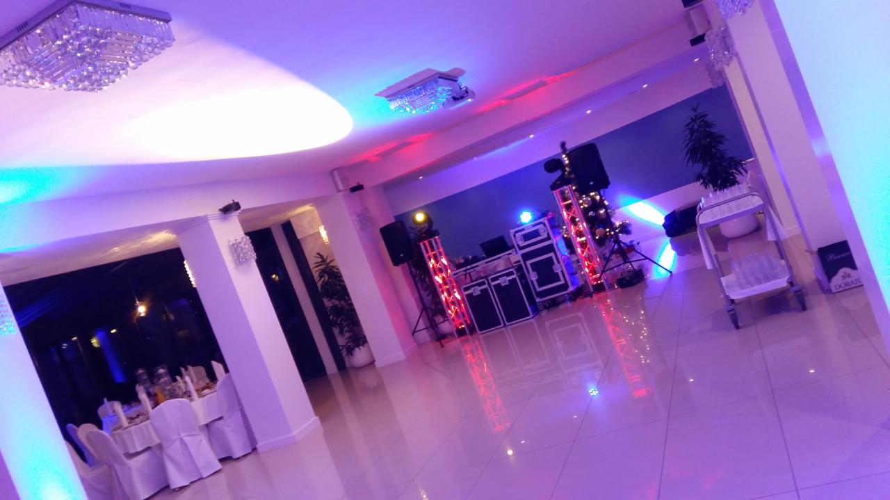 Sala bankietowa przygotowana na uroczystość, widoczne stoły z białymi obrusami, dekoracje świetlne i sprzęt nagłośnieniowy.