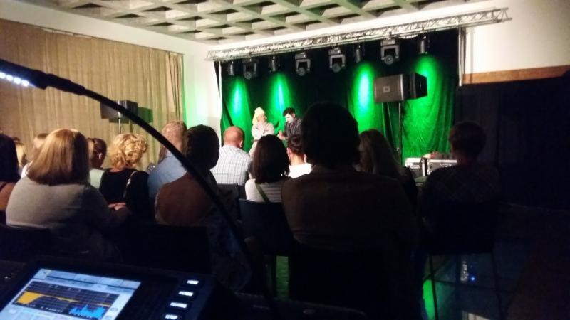 Widok zza konsolety dźwiękowej na scenę z zielonym oświetleniem i publicznością podczas występu.