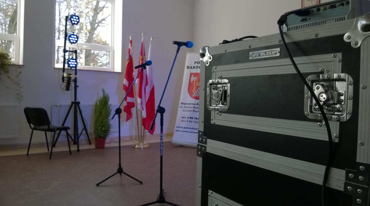 Przygotowane nagłośnienie i oświetlenie na sali z flagami, widoczny case na sprzęt marki Reloop oraz reflektor LED na statywie.