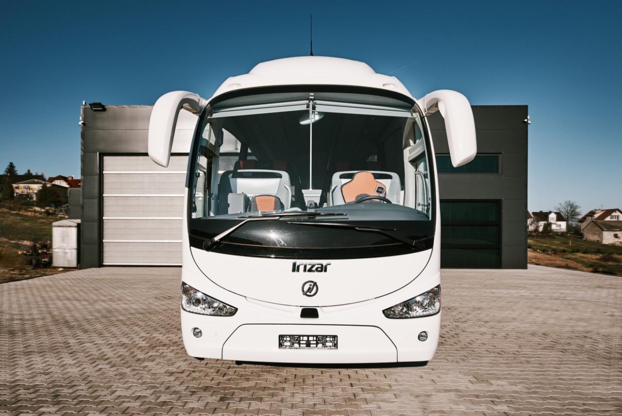 Biały autokar Irizar zaparkowany przed nowoczesnym budynkiem z szarą elewacją i bramą garażową, widok z przodu na tle błękitnego nieba.