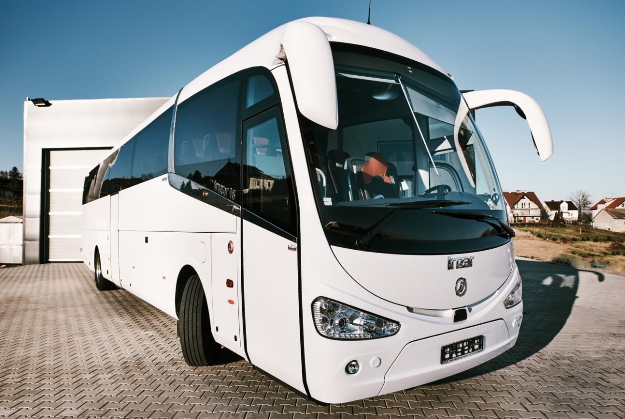 Nowoczesny, biały autokar turystyczny marki Irizar, zaparkowany na brukowanym placu przed budynkiem z otwartą bramą garażową w słoneczny dzień.