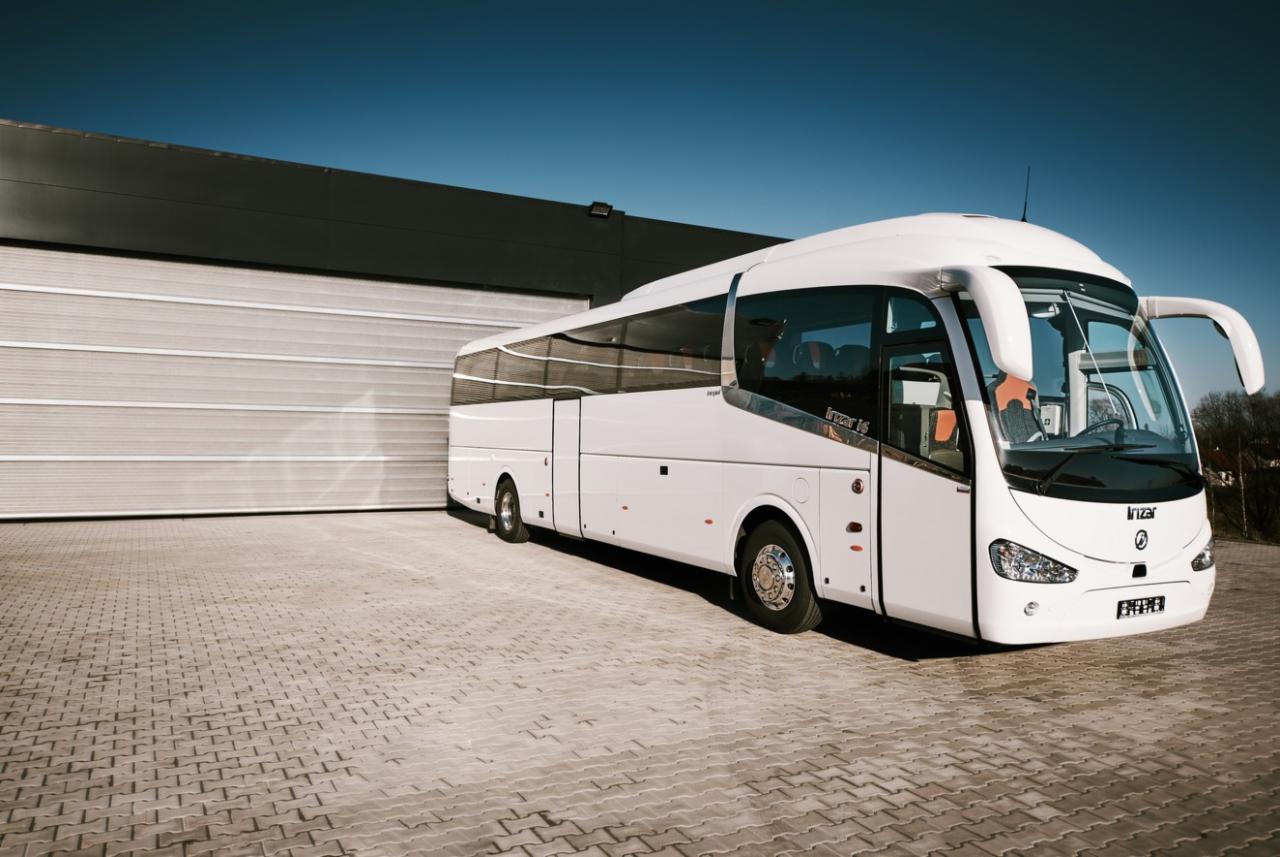 Biały autokar turystyczny marki Irizar zaparkowany przed szarą bramą garażową w słoneczny dzień.