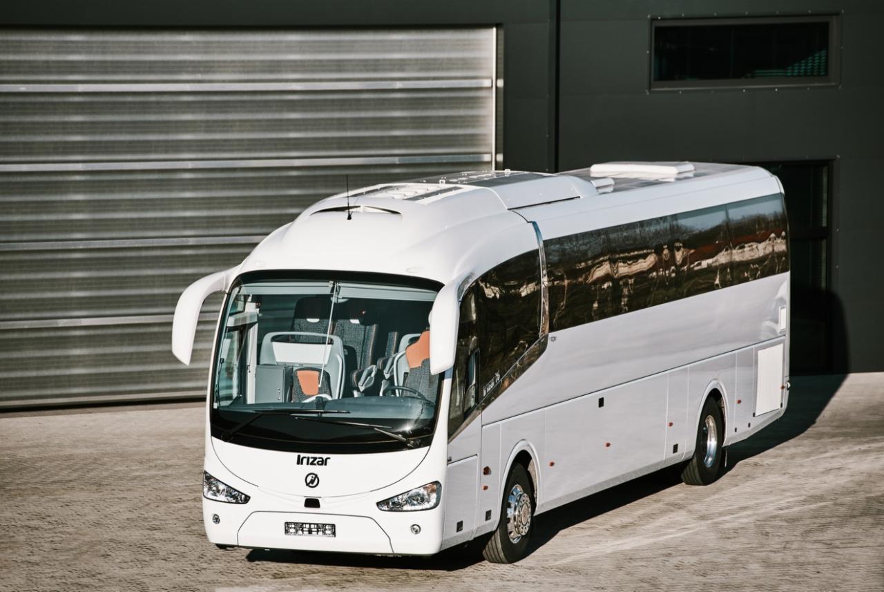 Nowoczesny, biały autokar Irizar z panoramicznymi oknami i reflektorem dachowym, zaparkowany przed szarą, metalową bramą garażową.