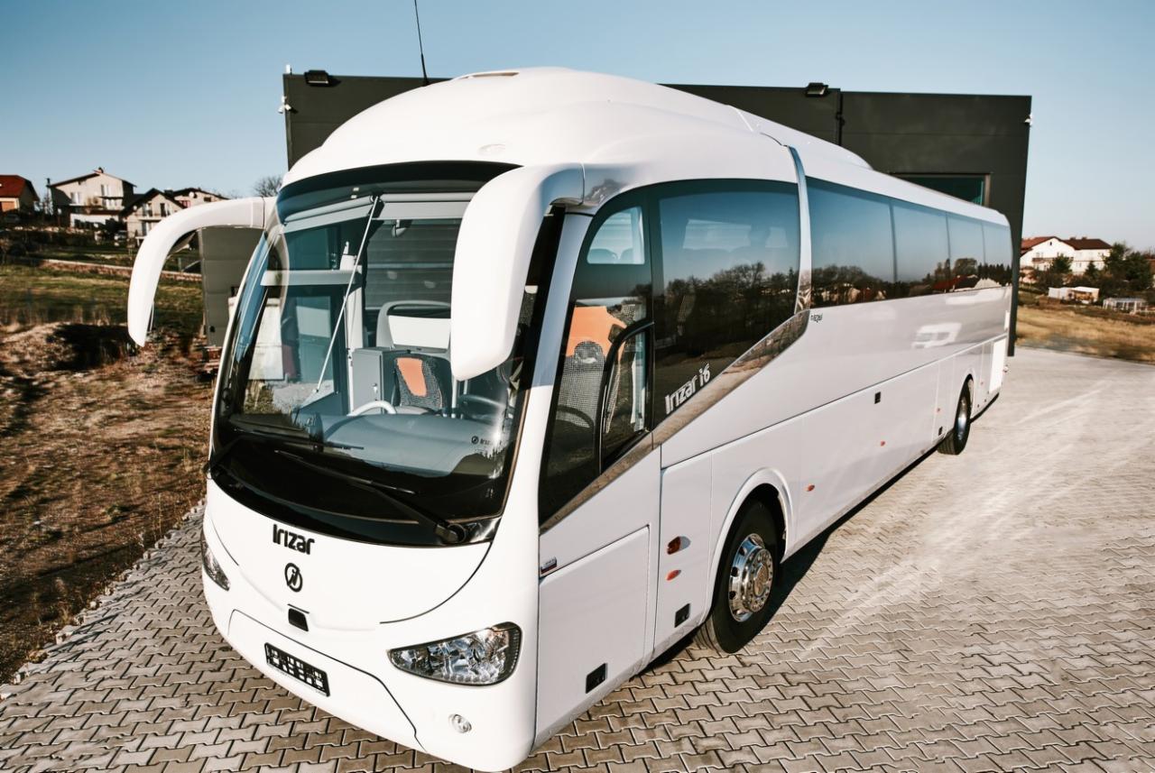 Nowoczesny, biały autokar Irizar i6 na brukowanym placu, z widocznym wnętrzem kabiny kierowcy i odbiciem zabudowań w szybach.