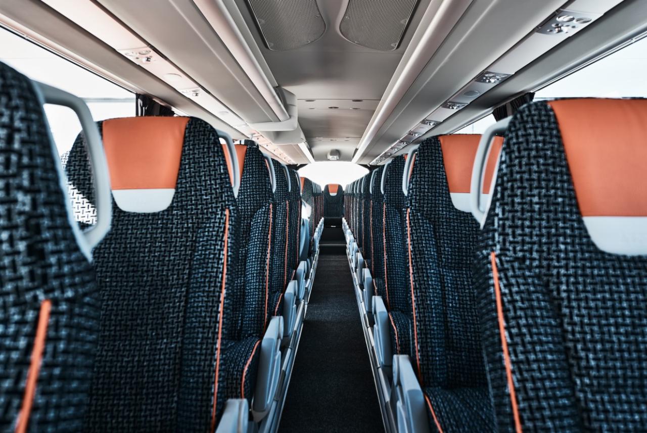 Wnętrze autobusu z rzędami foteli obitych wzorzystą tkaniną w odcieniach szarości i czerni, pomarańczowe zagłówki i jasne półki bagażowe nad głowami pasażerów.