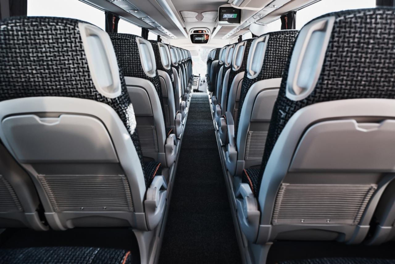 Wnętrze autobusu z rzędami szarych foteli z czarno-białą tapicerką, widok wzdłuż korytarza, ekrany multimedialne zamontowane pod sufitem.