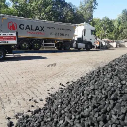 Ciężar&oacute;wka firmy Galax z napisem 'Eksperci od ekogroszku' na tle sterty czarnego węgla na placu składowym.