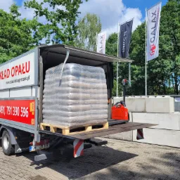 Ciężarówka z otwartą platformą załadunkową, na której znajduje się paleta z workami opału, całość owinięta folią. W tle widać wózek widłowy, betonowe bloki, drzewa i flagi z logo firmy.