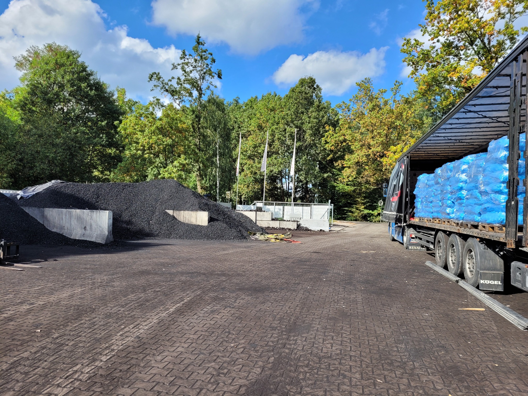 Kopalnia odkrywkowa z hałdami czarnego węgla, zaparkowaną ciężarówką z niebieskimi workami i zielonymi drzewami w tle w słoneczny dzień.