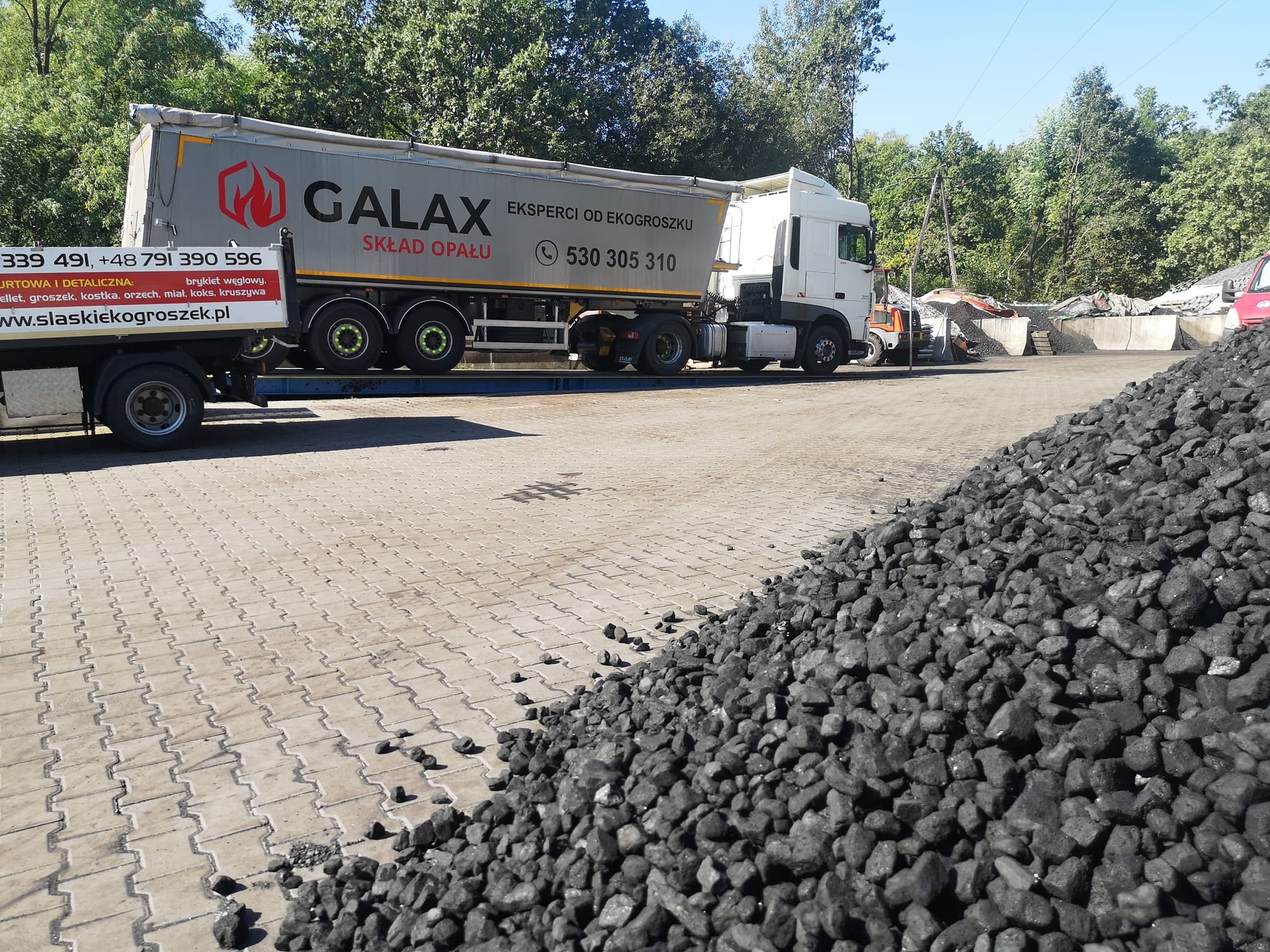 Ciężarówka firmy Galax z napisem 'Eksperci od ekogroszku' na tle sterty czarnego węgla na placu składowym.