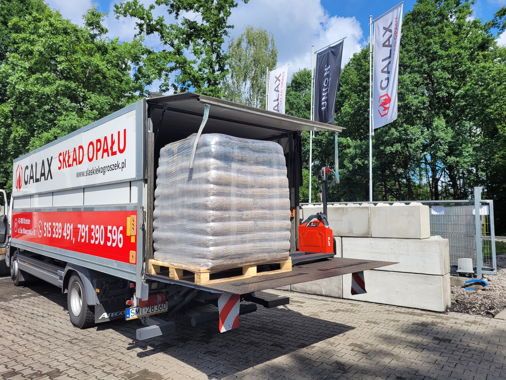 Ciężarówka z otwartą platformą załadunkową, na której znajduje się paleta z workami opału, całość owinięta folią. W tle widać wózek widłowy, betonowe bloki, drzewa i flagi z logo firmy.