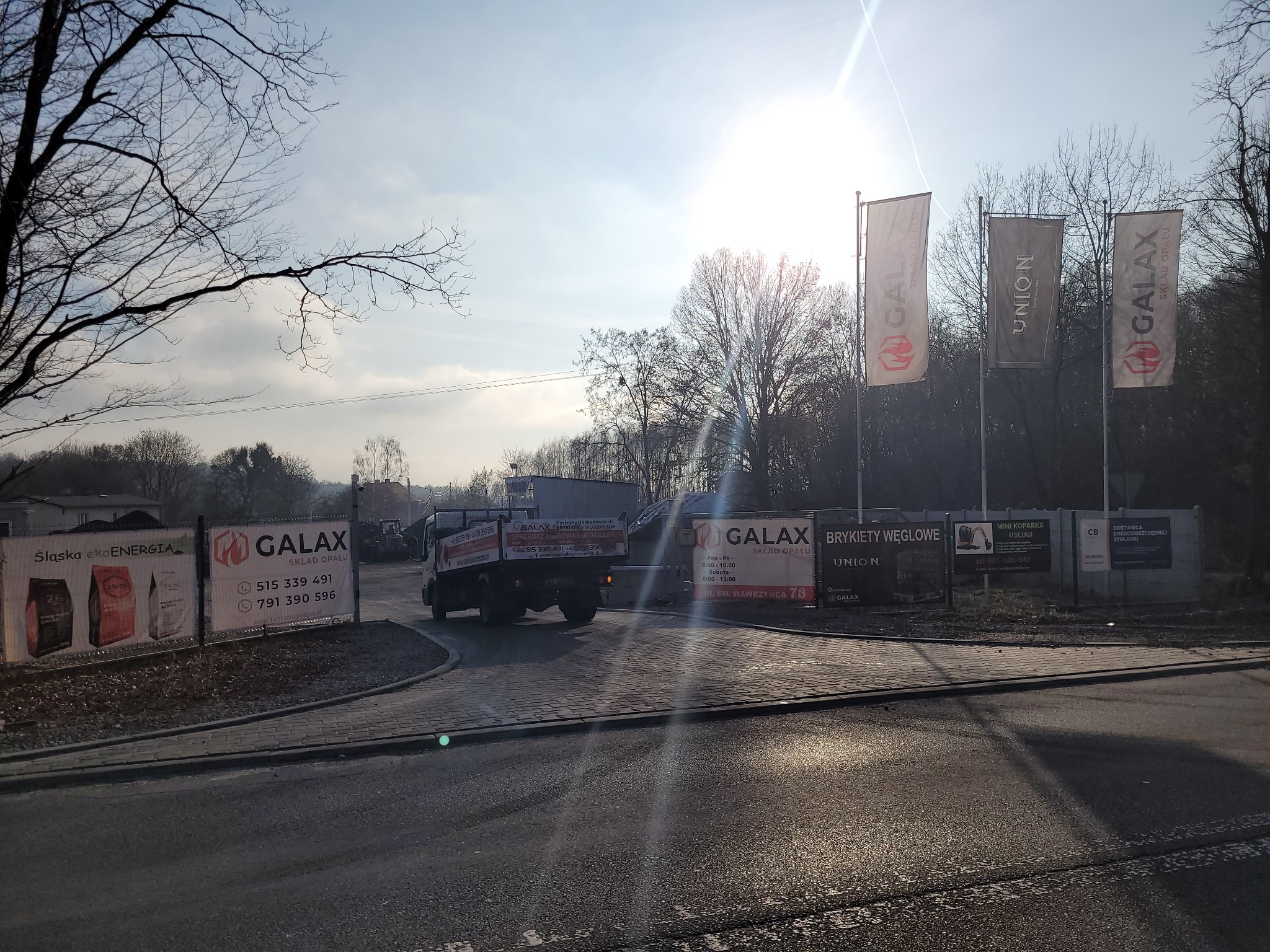 Ciężarówka wjeżdża na teren składu opału Galax, widoczne banery reklamowe ekogroszku i brykietów węglowych, flagi z logo firmy i nazwa ulicy Św. Wawrzyńca 78.