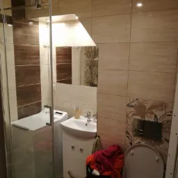 Wykończona łazienka z kabiną prysznicową, pralką obok umywalki i beżowo-brązowymi płytkami na ścianach, widoczne akcenty dekoracyjne w postaci kwiatowych wzorów.