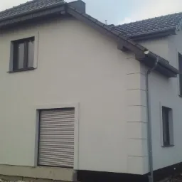 Dwupiętrowy dom z jasną elewacją, szarą dachówką i brązowymi elementami wykończeniowymi okien i dachu, widoczny z boku w pochmurny dzień.