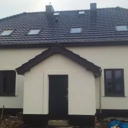 Otynkowany dom jednorodzinny z ciemną podmurówką, stolarką okienną i drzwiami, widoczne okna połaciowe w ciemnym dachu z dachówki ceramicznej, teren wokół domu częściowo uporządkowany.